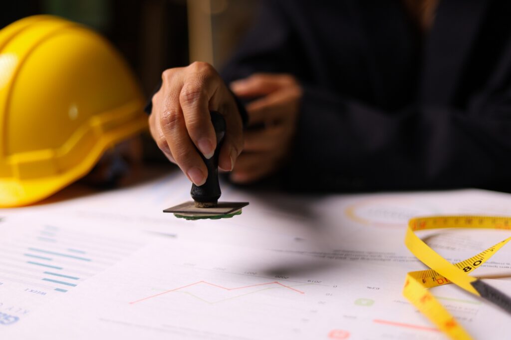 Construction BluePrints On A Table With A Helmet In The Background. A hand holds an approval stamp and is about to stamp the blue prints.